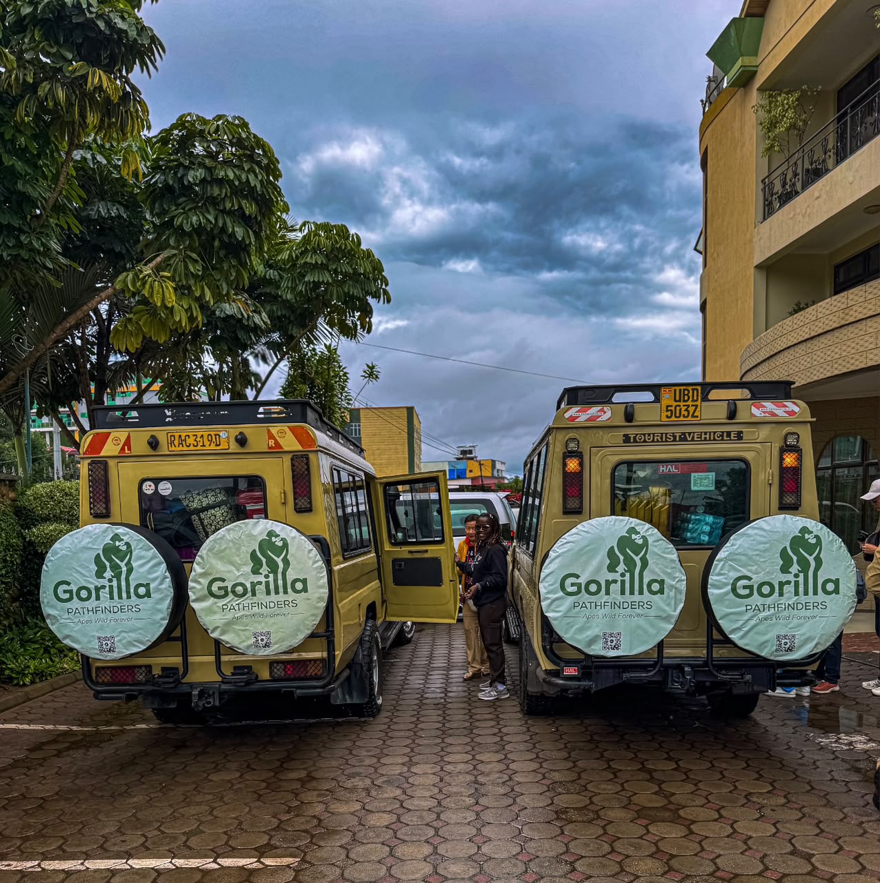 Travellers standing between two beige Gorilla Pathfinders safari vehicles outside a lodge
