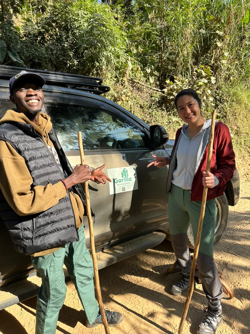 Smiling traveller and safari guide holding wooden trekking sticks beside a grey vehicle on a forest road