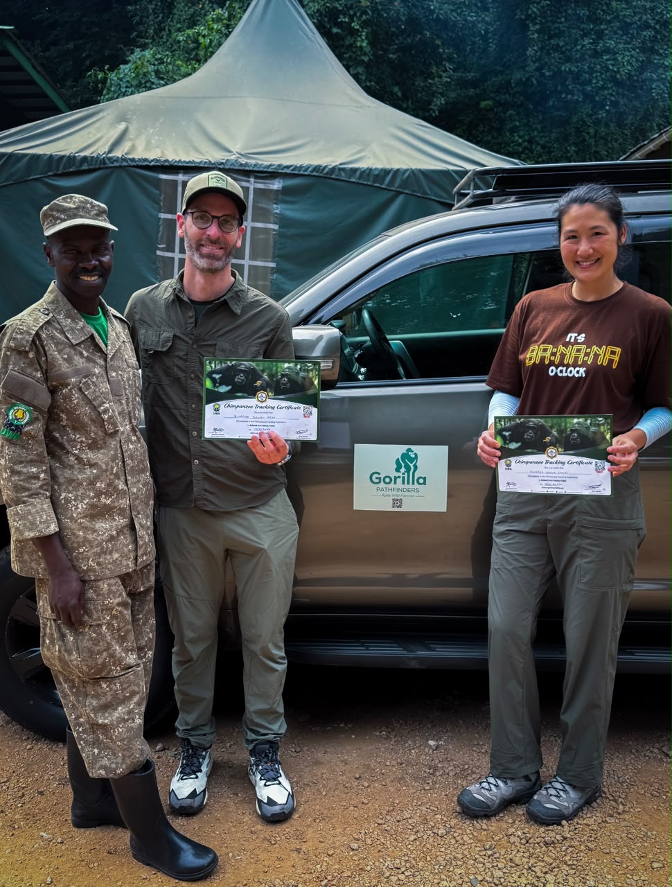 Two travellers and a ranger holding chimpanzee tracking certificates beside a safari vehicle