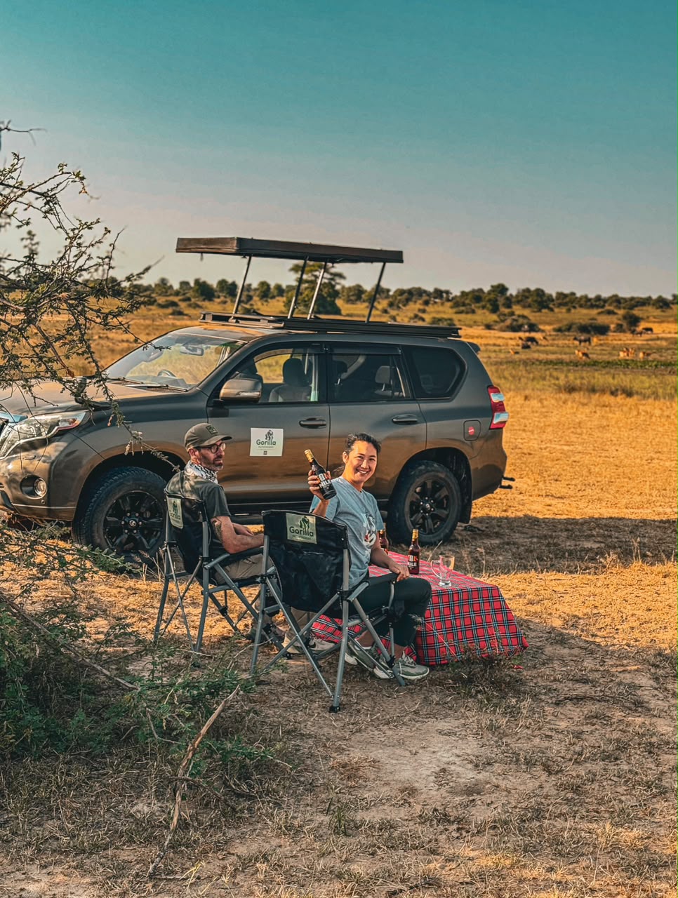 Two safari guests relaxing at a picnic beside a grey vehicle in the open savannah