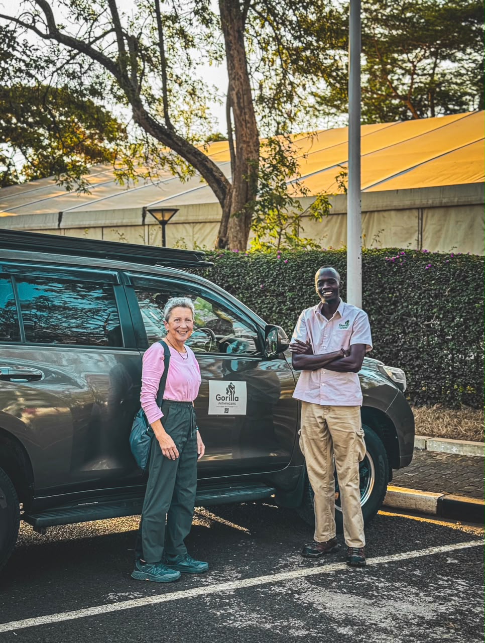 Smiling woman traveller and Gorilla Pathfinders guide standing beside a grey safari vehicle