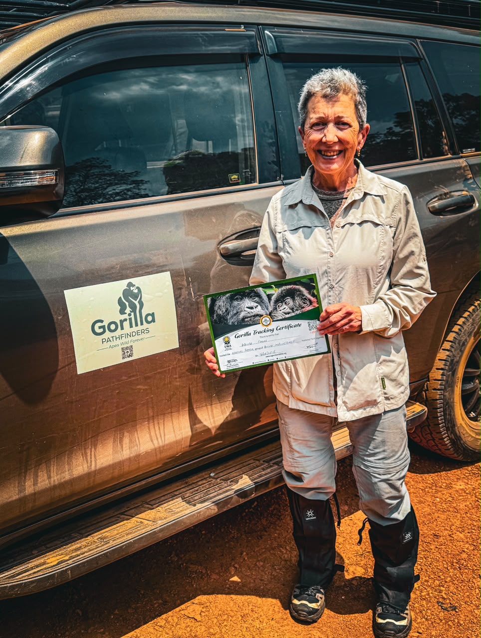 Smiling woman holding a gorilla tracking certificate beside a mud-splashed safari vehicle
