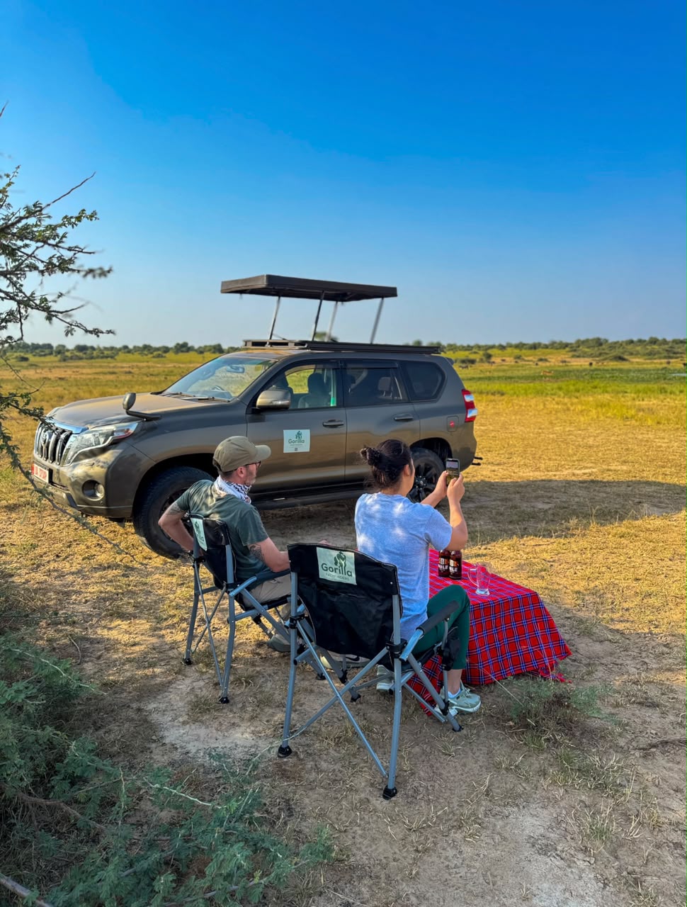 Two safari guests seated at a picnic table beside a pop-up-roof vehicle in open savannah